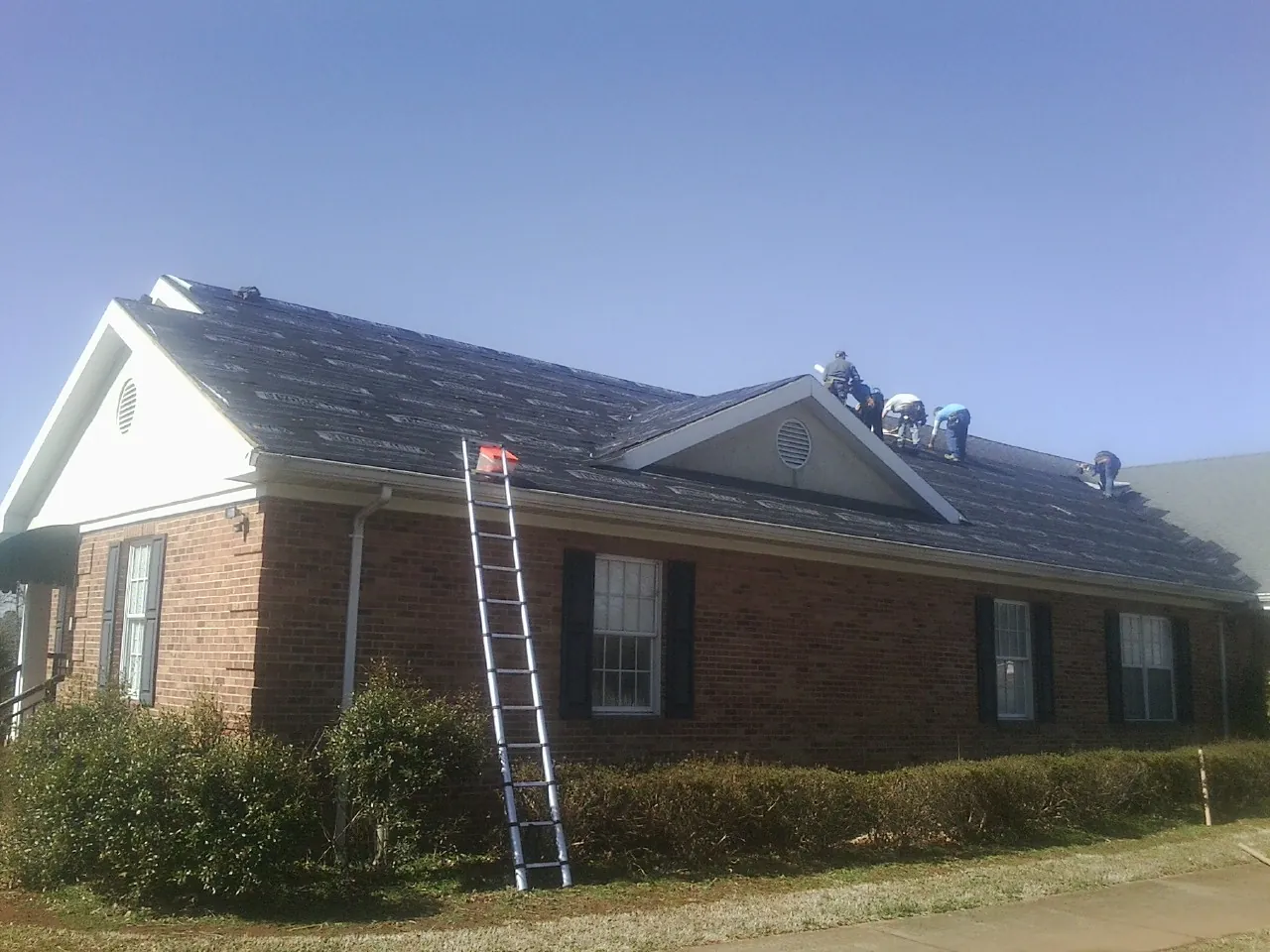 Crew tearing shingles off a brick ranch roof with a ladder set — Metal Roof Repair work by our crew in Lomira