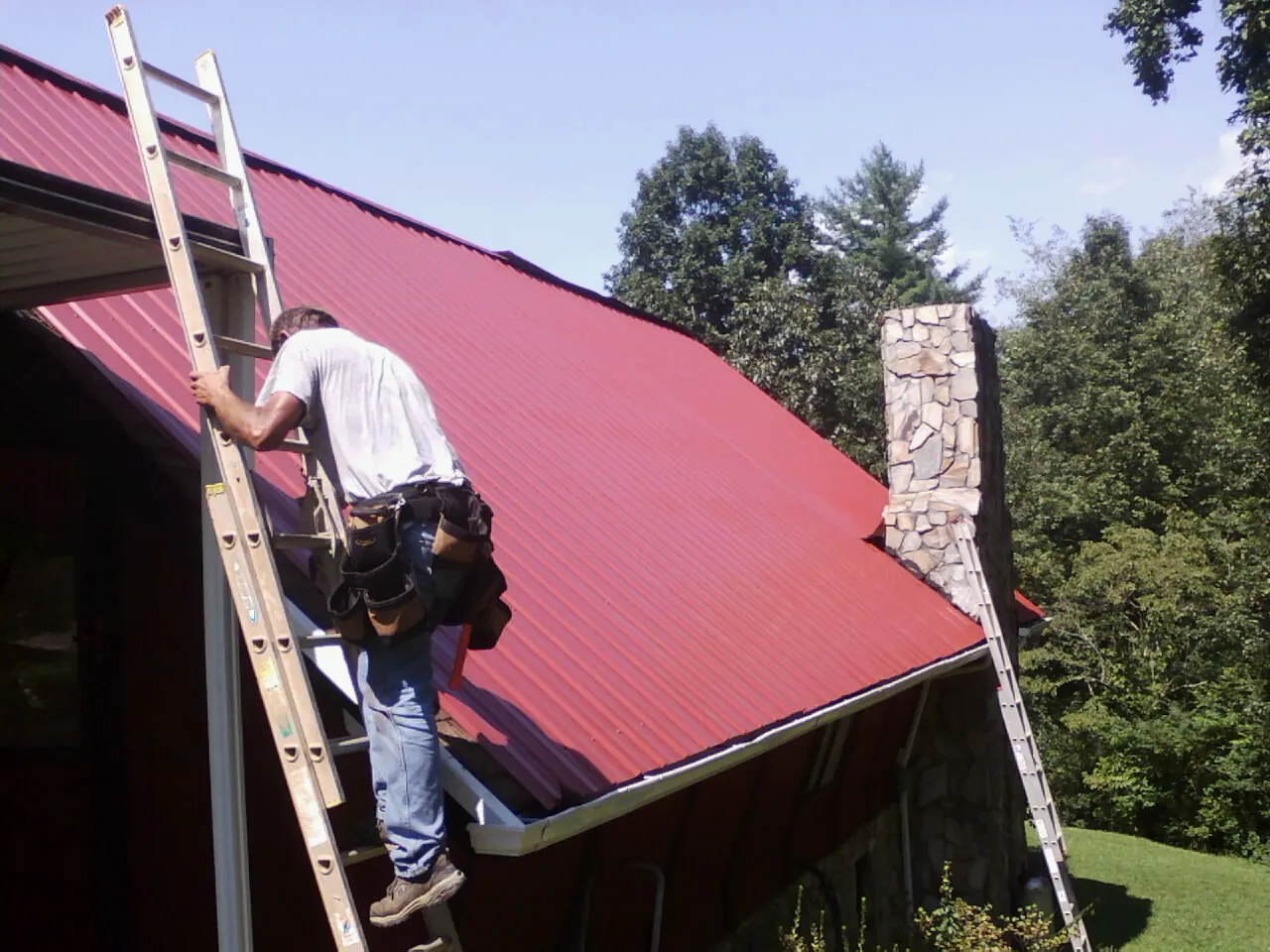Roofer on a ladder beside a red metal roof and stone chimney — Soffit & Fascia Repair by Roofing Brothers in Lomira